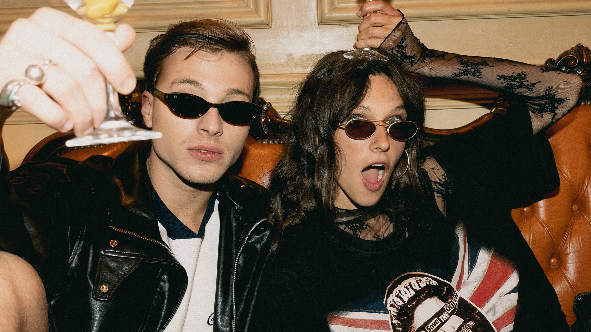 FORK Eyewear lifestyle scene, two friends raise a glass on a vintage sofa, narrow tortoise rectangle and small gold oval sunglasses.
